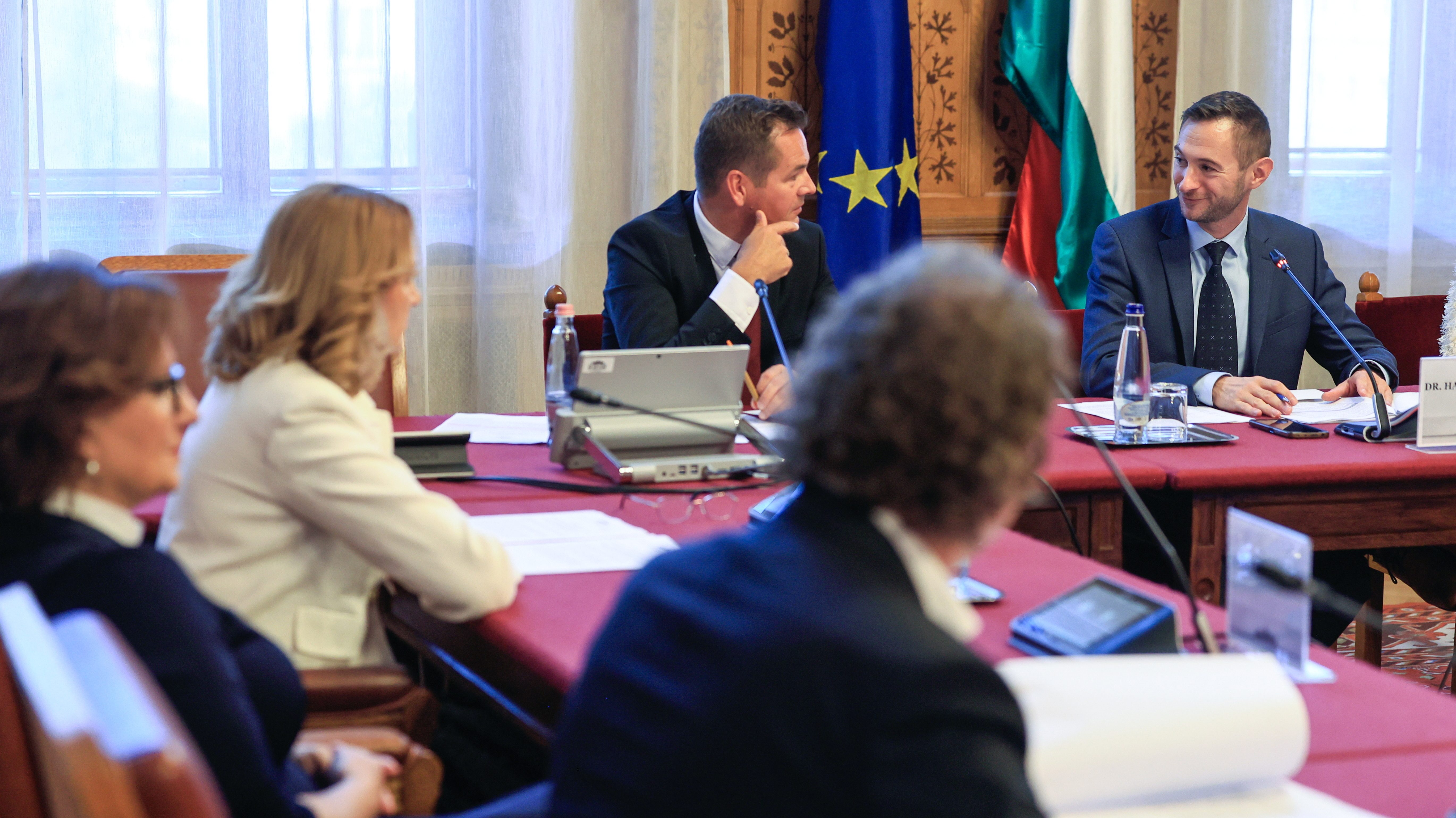 Professional meeting around a red table; microphones, papers, and water bottles visible, flags in the background