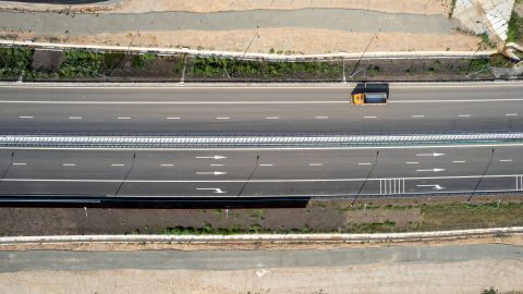Aerial view of a multi-lane highway with a yellow-orange truck driving in the right lane; grassy median and dirt shoulders on both sides.