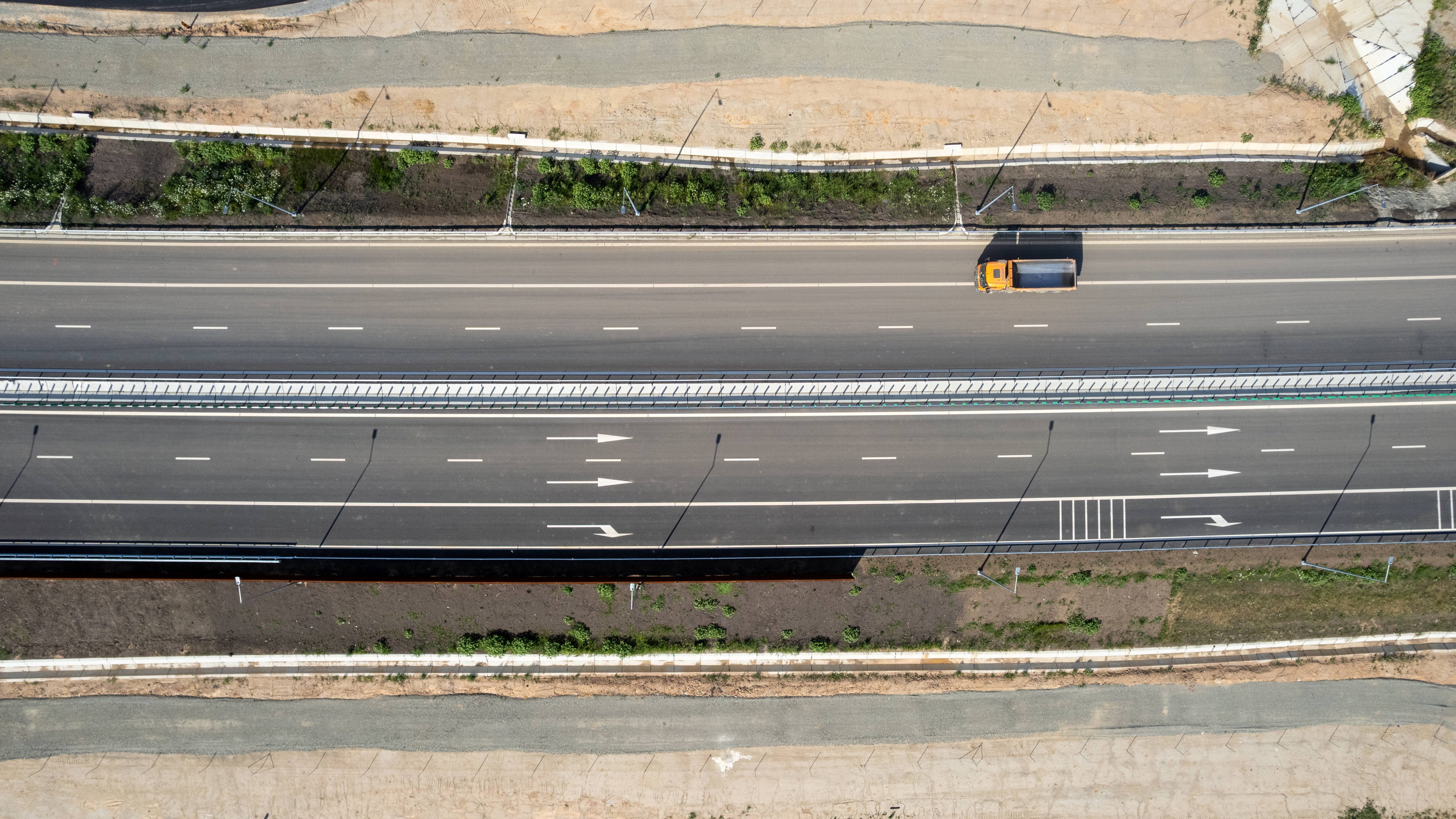 Aerial view of a multi-lane highway with a yellow-orange truck driving in the right lane; grassy median and dirt shoulders on both sides.