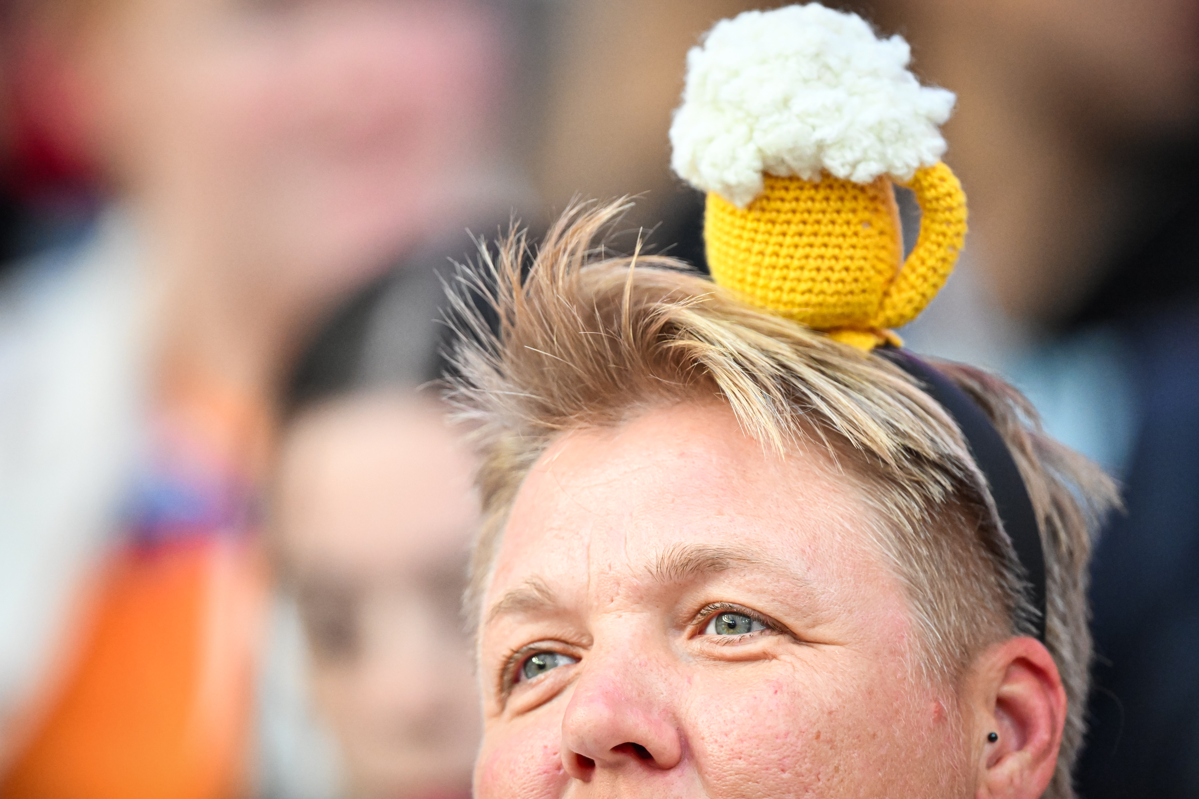 Close-up of a person wearing a yellow crochet beer mug hat with white foam on top, showing a green eye and part of the face.