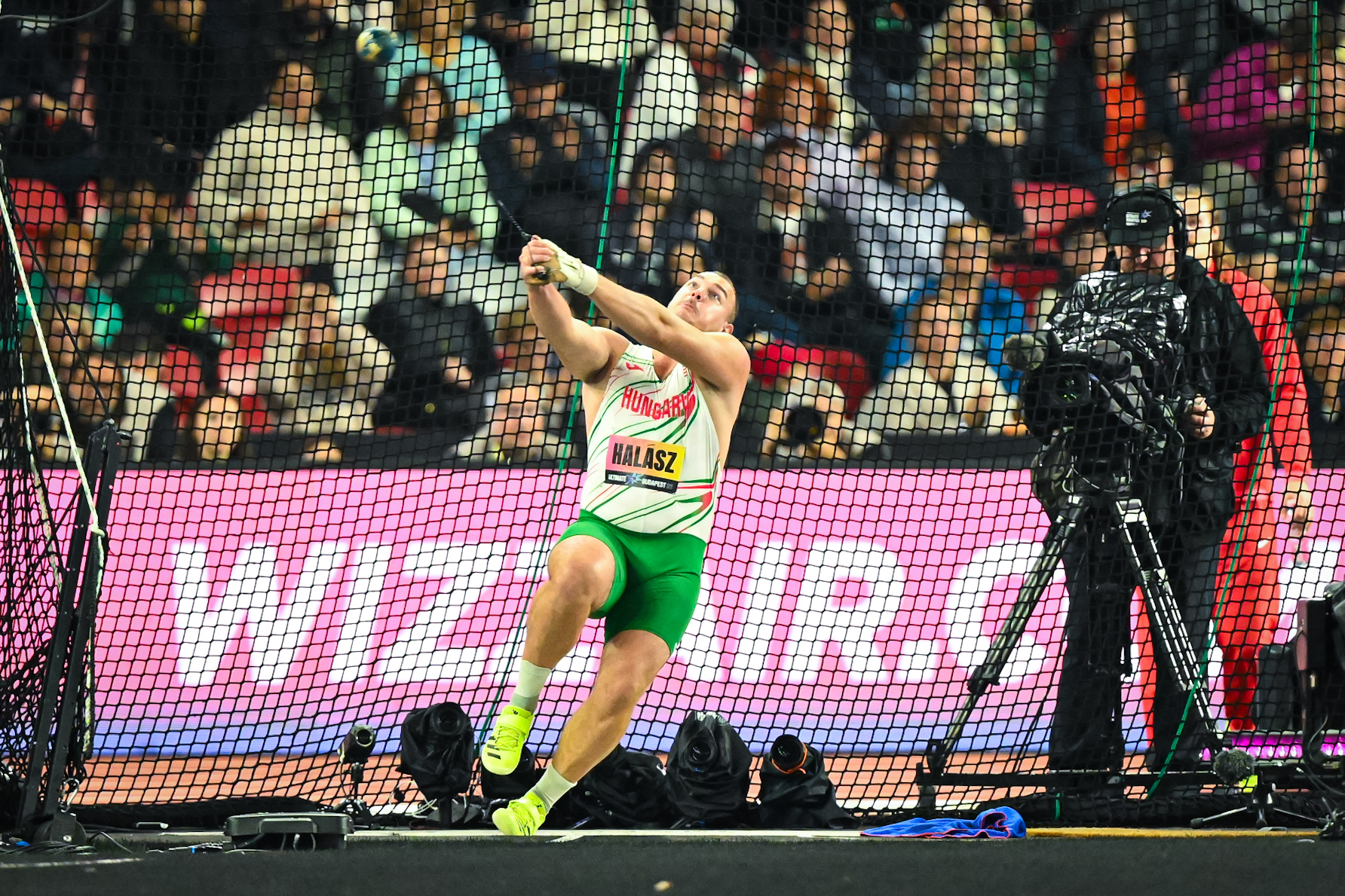 Hungarian male athlete in green shorts and white jersey reaching to strike a ball behind a protective net, crowd watching in the stands behind him.