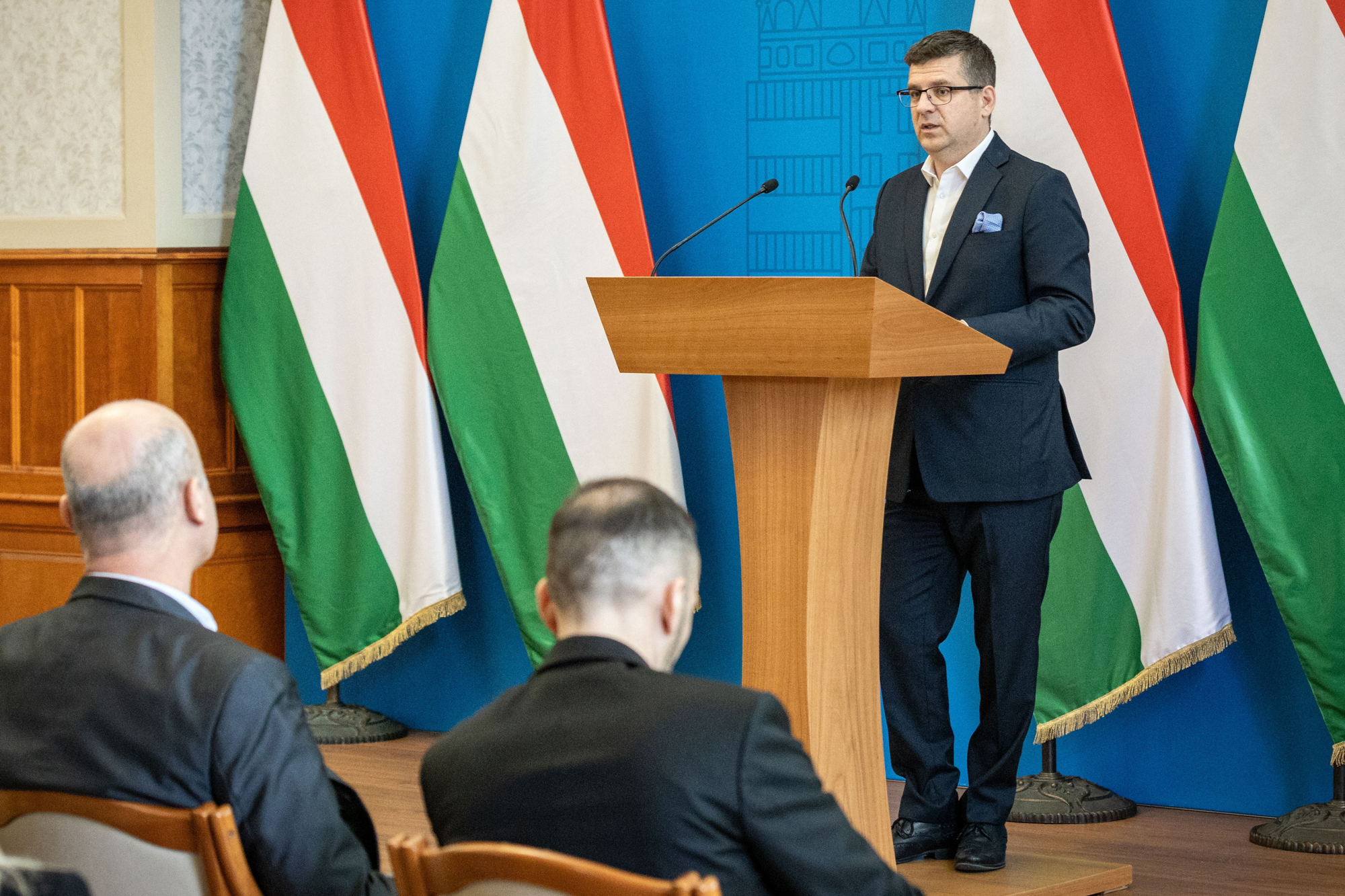 Man in a dark suit speaks at a wooden podium with microphones, colorful flags behind him.