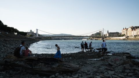 People explore a rocky riverbank by a wide river with a white suspension bridge and a cityscape in the background.