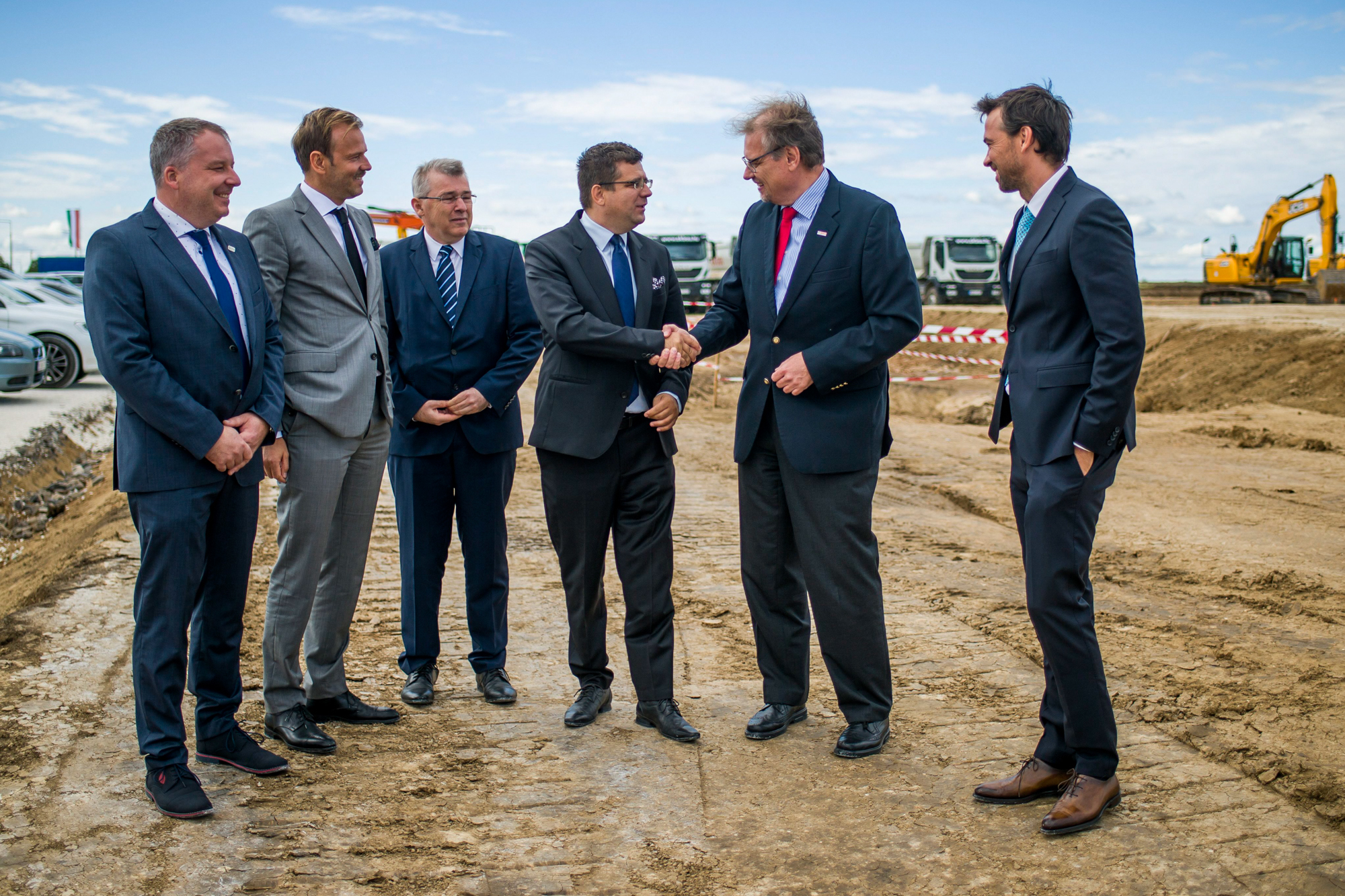 Five men in business suits stand on a dirt construction site, two of them shaking hands and smiling.