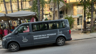 Dark blue van with 'Magyar Falu Program Zselickisfalud' logo parked on a city street; market stalls and trees visible in the background.