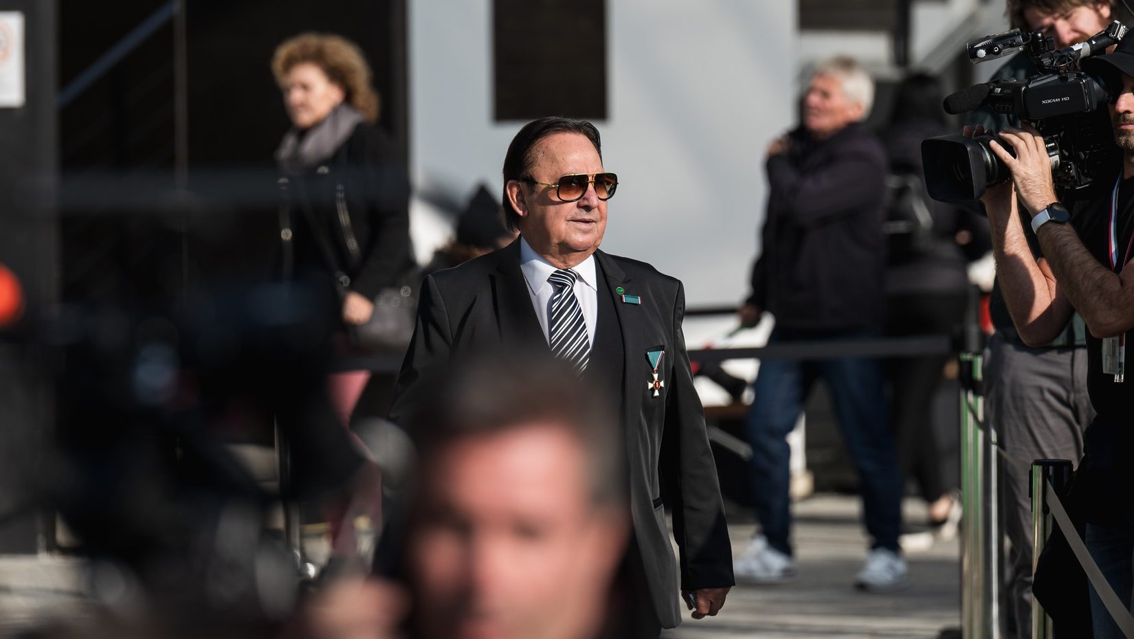 Man in a black suit with striped tie and medals walking past photographers at an outdoor event.