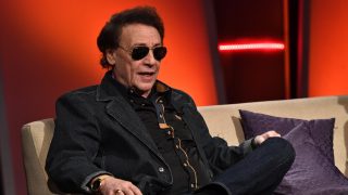 Man in a dark denim jacket and sunglasses seated on a beige couch against an orange stage backdrop, speaking or listening attentively.
