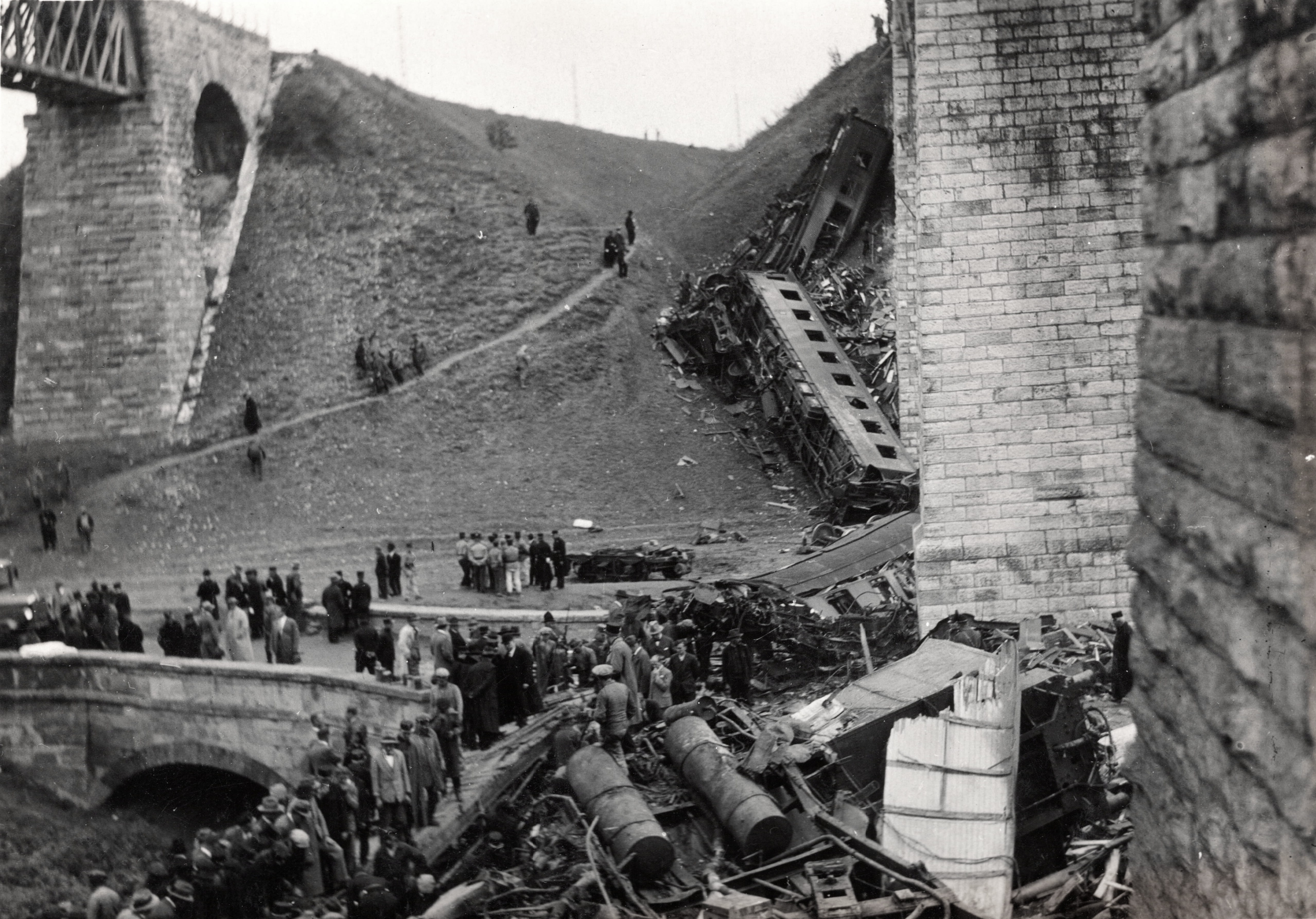 Crowd of people gathered around a collapsed railway or bridge wreckage on a hillside, with scattered metal and wood debris and a brick wall nearby.