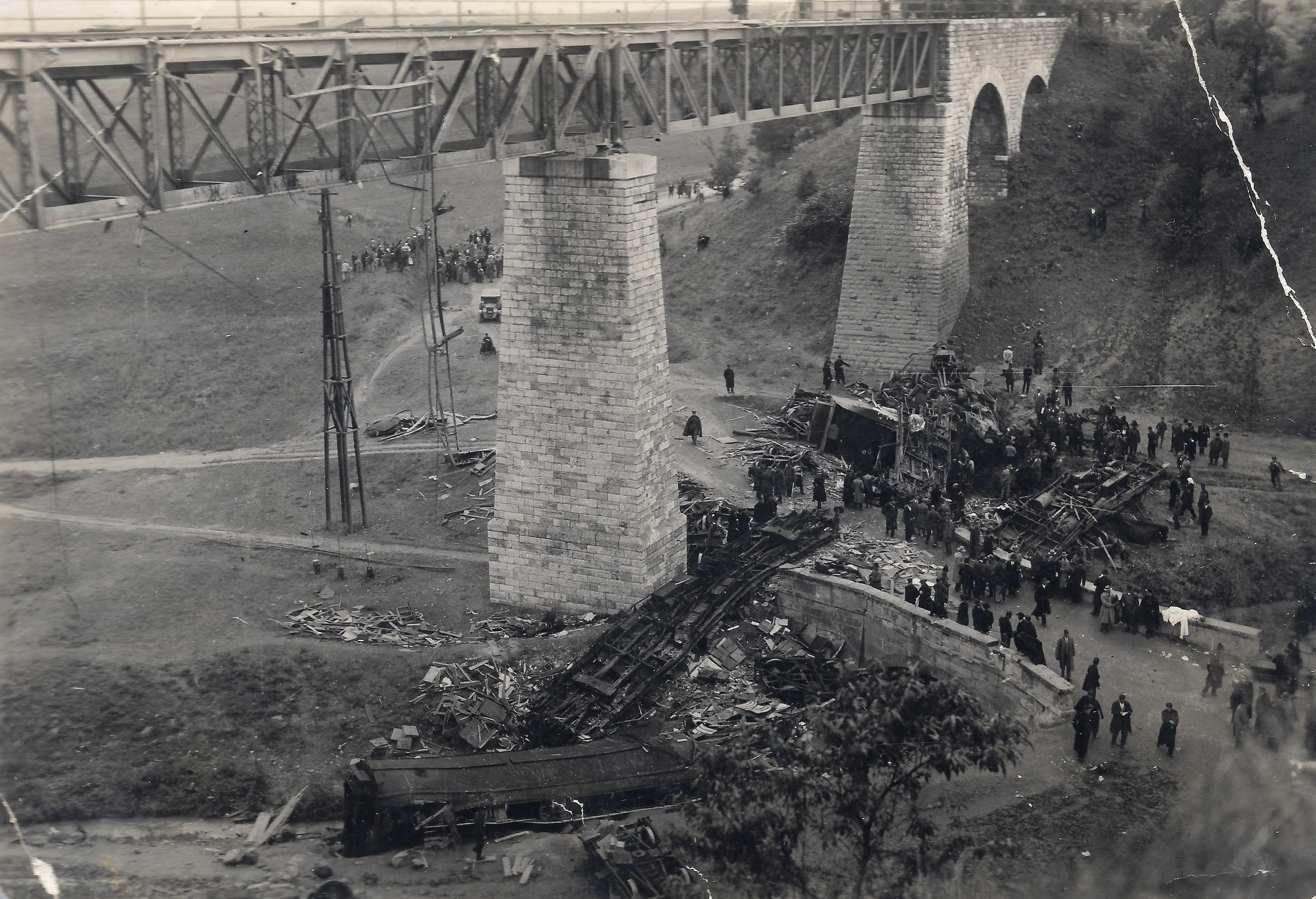 Historic black-and-white scene of a broken stone arch bridge with a collapsed railway line and a crowd inspecting wreckage beneath.