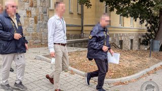 Three people walk along a brick building; two wear police jackets, one in a white shirt carries a badge and a lanyard, while another officer holds papers.