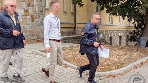 Three people walk along a brick building; two wear police jackets, one in a white shirt carries a badge and a lanyard, while another officer holds papers.