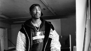 Black-and-white portrait of a man wearing a backwards cap, chains, and a patterned jacket in a dim hallway.