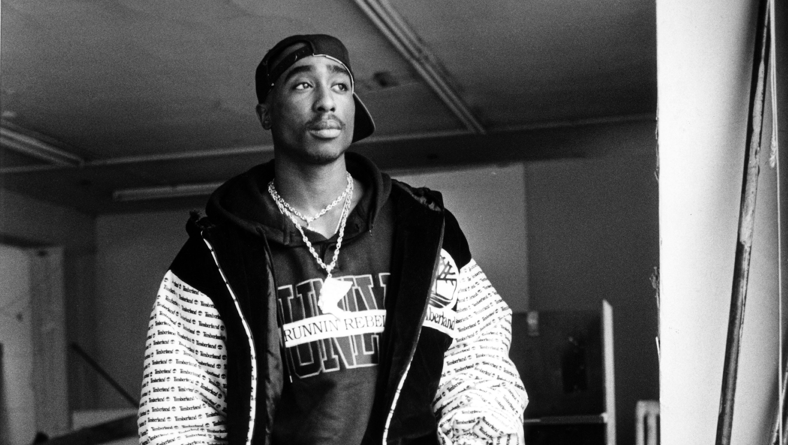 Black-and-white portrait of a man wearing a backwards cap, chains, and a patterned jacket in a dim hallway.