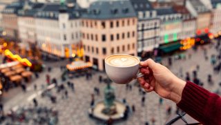 Close-up of a hand in a red sleeve holding a cup of latte with a heart-shaped latte art, overlooking a blurred city square.