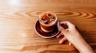 Hand holding a glass of iced coffee with ice cubes on a wooden table.