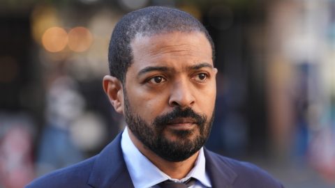Close-up of a man with a short beard in a navy suit and light blue shirt, looking to the right outdoors.