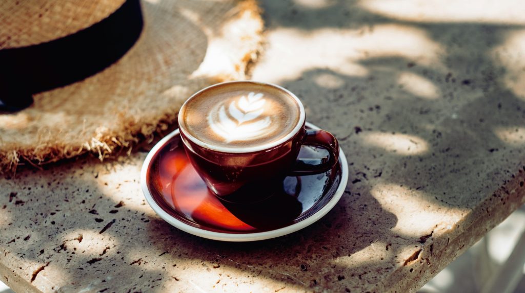 Brown ceramic cup of latte with leaf latte art on a matching saucer, set on a sunlit, textured stone surface with a straw hat in the background.