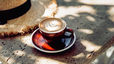 Brown ceramic cup of latte with leaf latte art on a matching saucer, set on a sunlit, textured stone surface with a straw hat in the background.