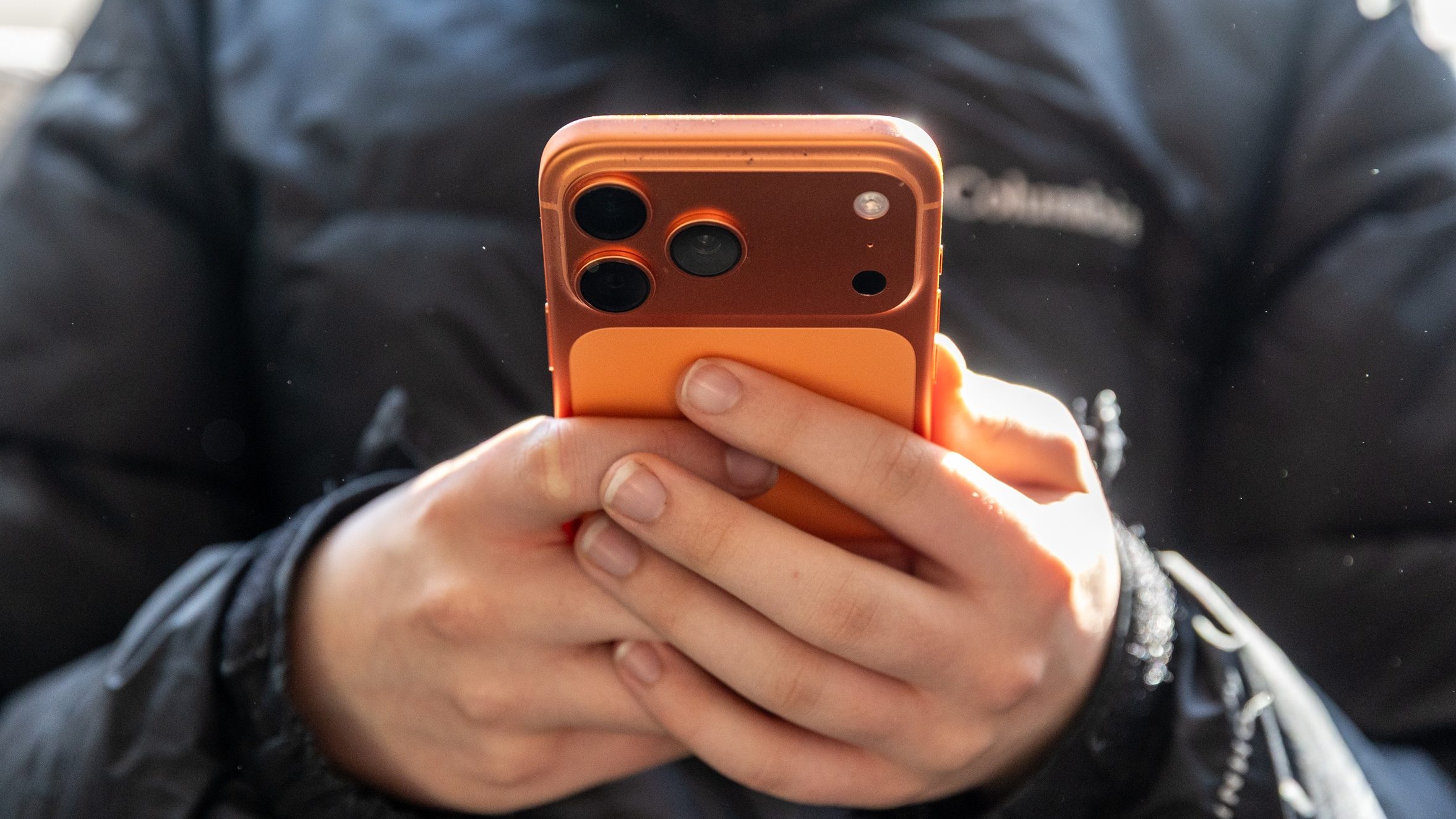 Person wearing a dark jacket holding an orange iPhone with a triple-camera back, close-up of hands.