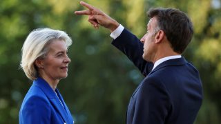 Two formally dressed people outdoors: a man in a dark suit points toward the distance while facing a woman in a blue blazer.
