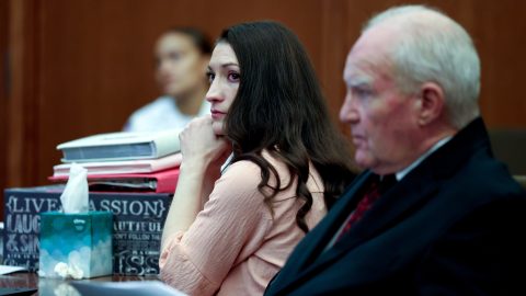 Woman with long dark hair resting her chin on her hands, seated beside an older man in a suit during a meeting; papers and tissues on the table nearby.