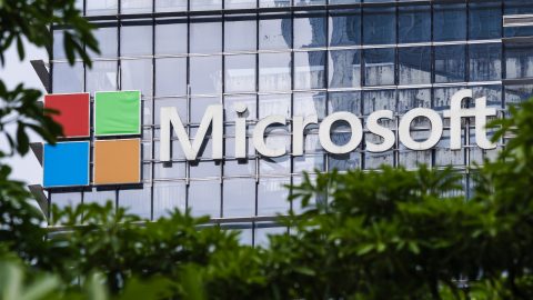 Microsoft logo displayed on a glass office building with colorful square panels on the left, partially framed by greenery