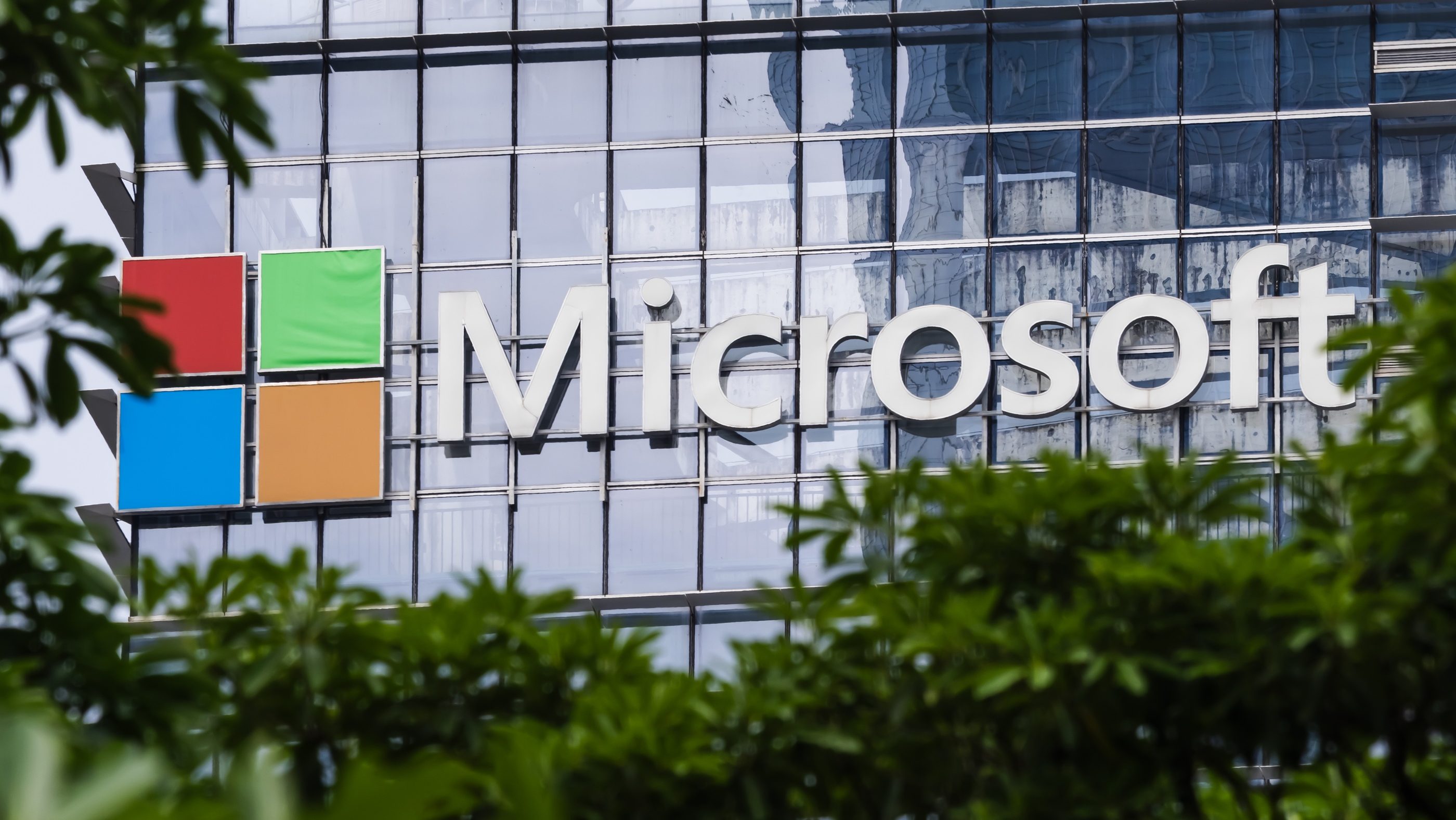 Microsoft logo displayed on a glass office building with colorful square panels on the left, partially framed by greenery