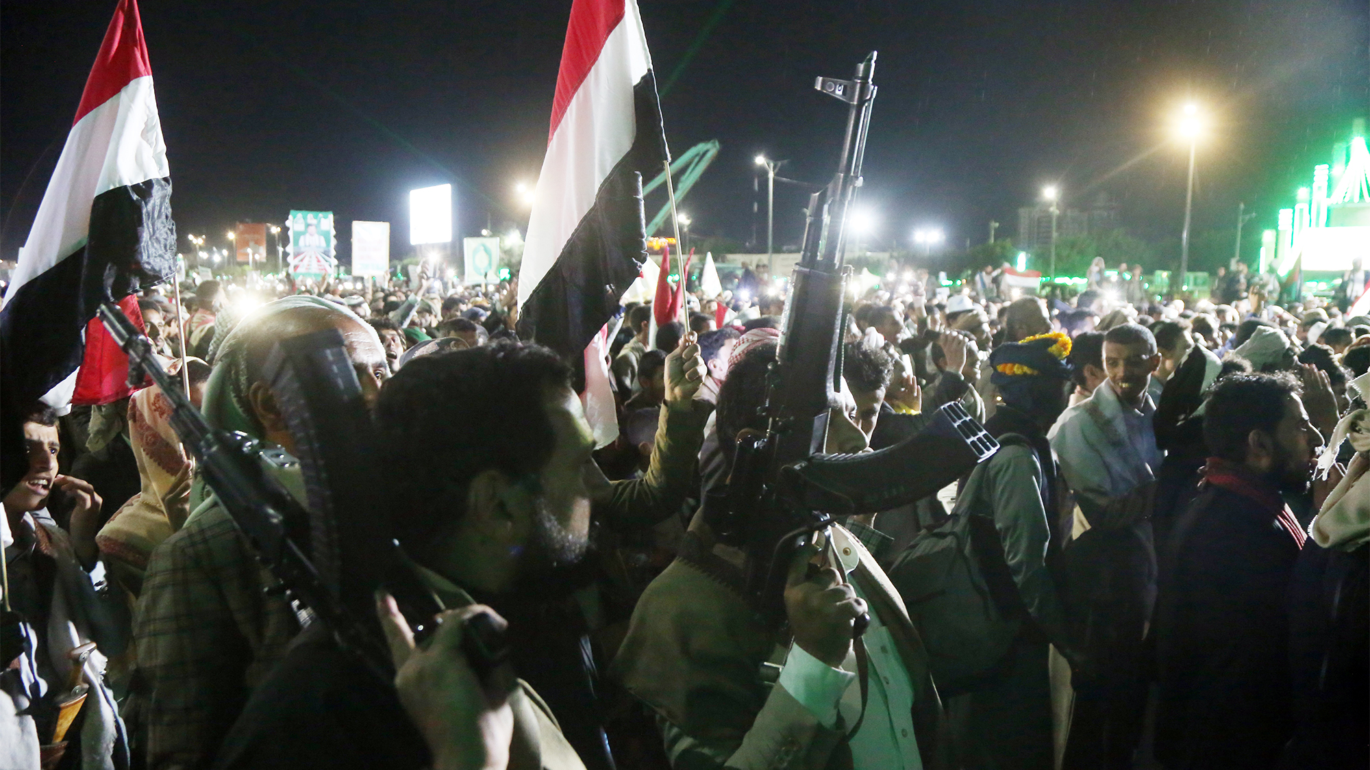 Large crowd at night holding red-white-black flags in what appears to be a protest or rally, with some people carrying rifles raised above the crowd.