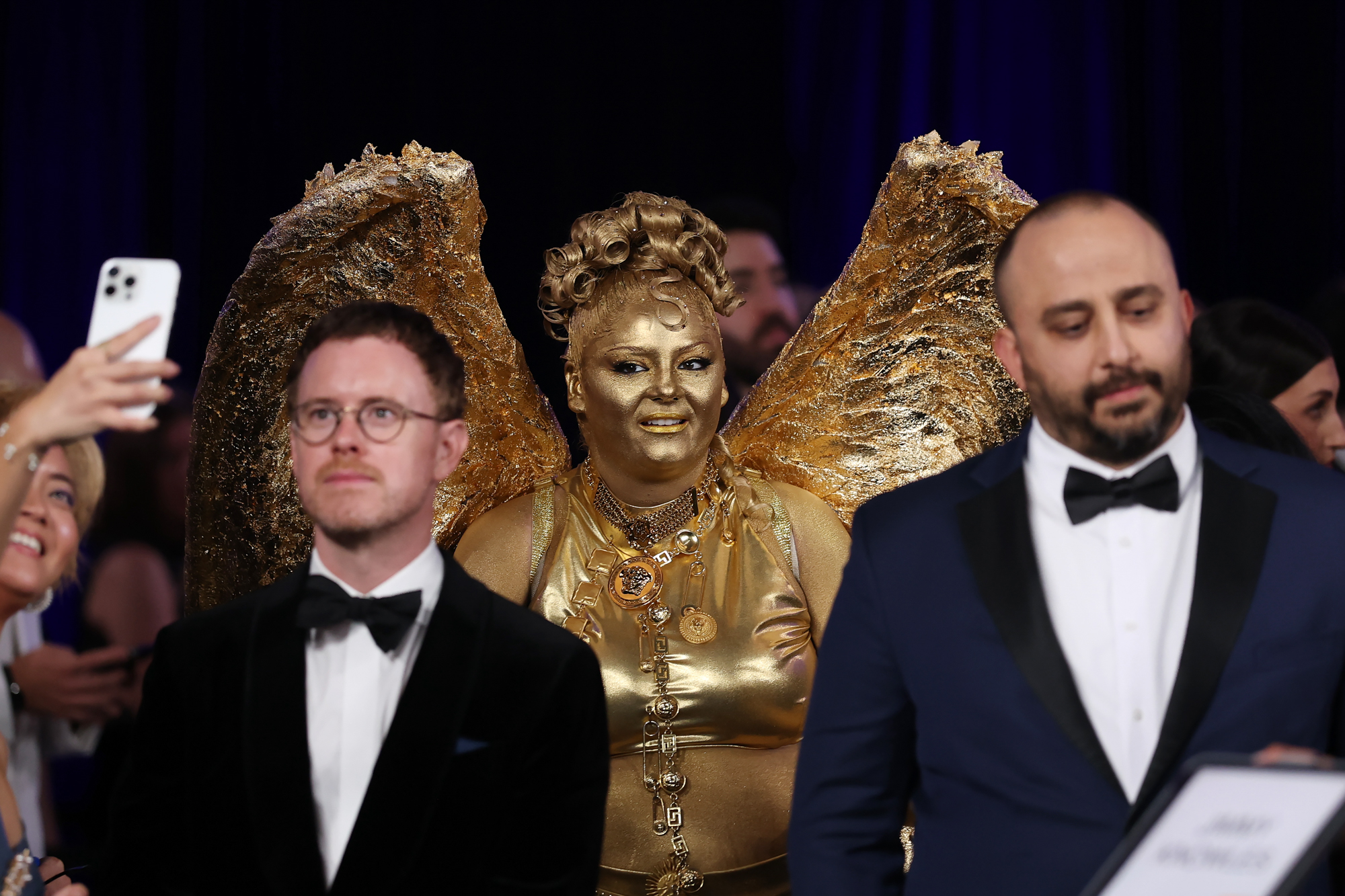 Person in gold body paint and elaborate costume with large gold wings at a formal event among suited guests.
