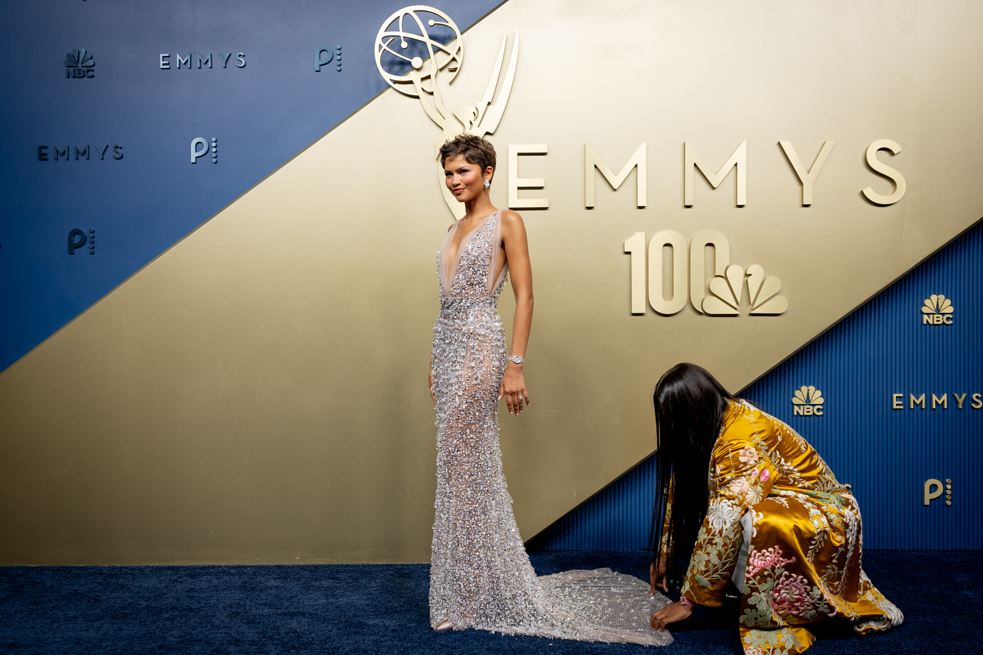 Woman in a sparkling silver gown poses on the Emmy Awards backdrop with a kneeling person in the foreground amid the red-carpet setup.