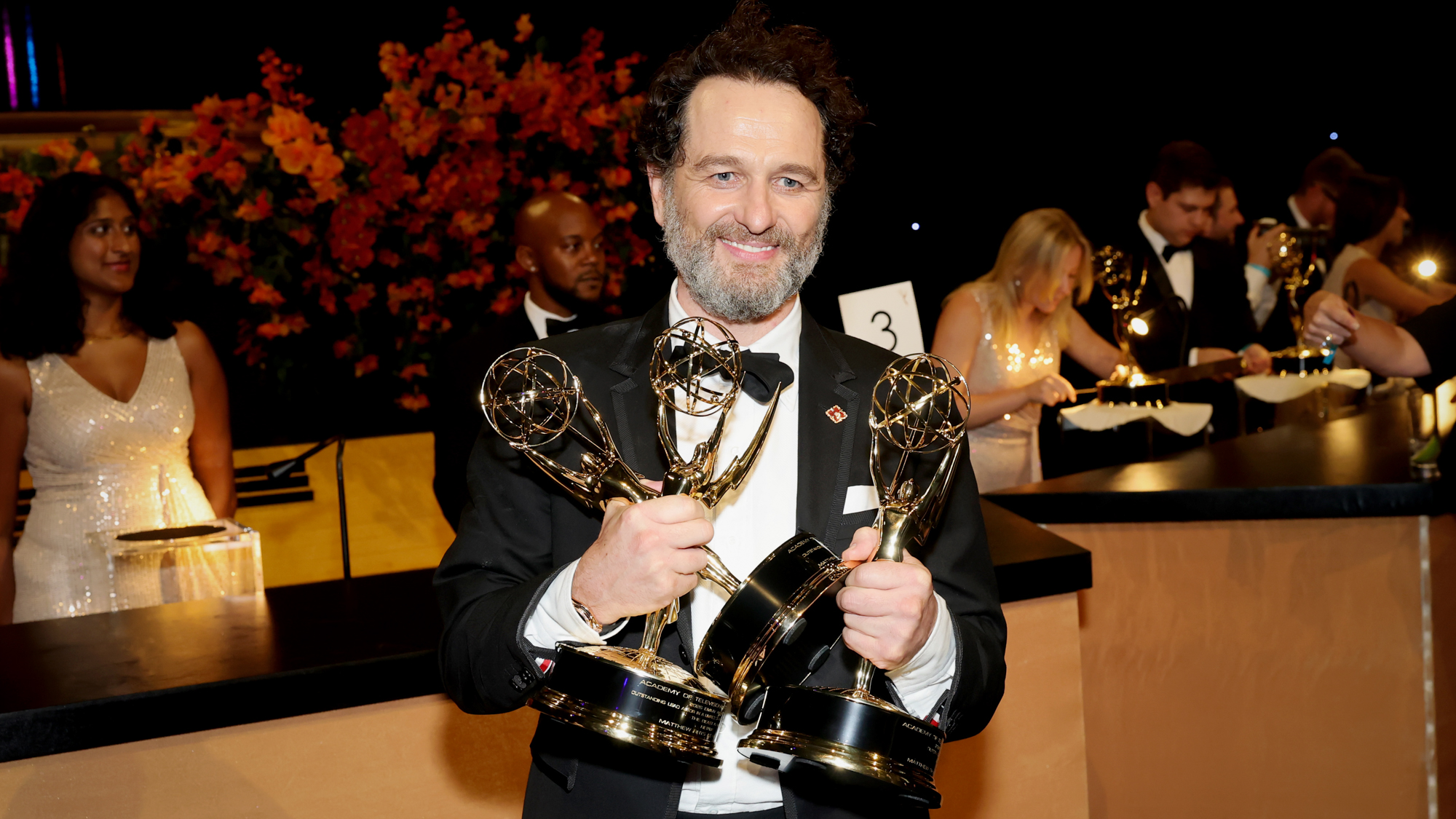Smiling man in a black tuxedo holding three gold trophy awards at a formal gala, with orange flowers in the background.