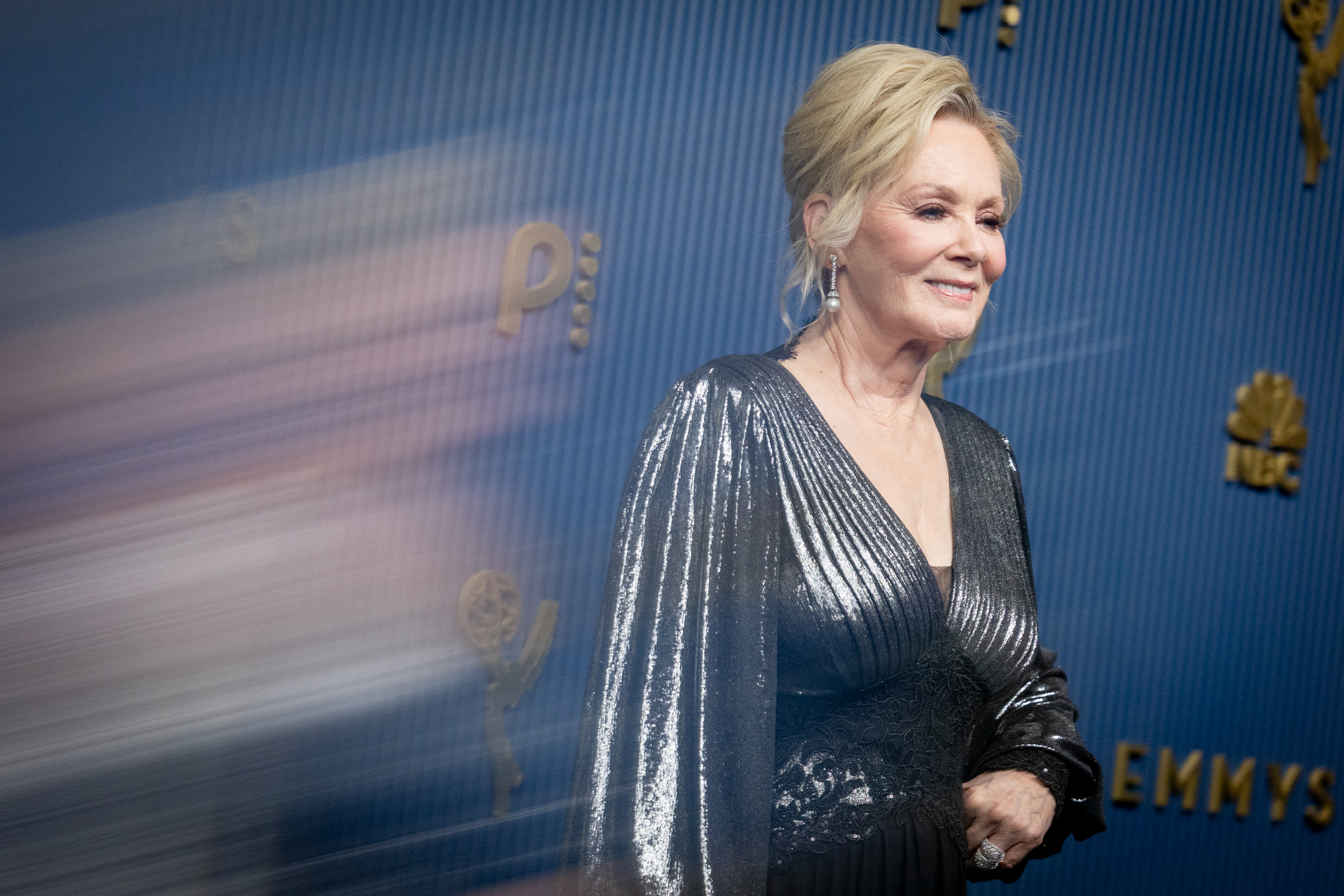 Smiling woman in a silver sequin gown posing on a blue Emmy Awards backdrop.