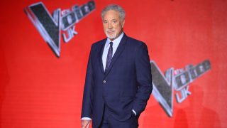 Older man in a dark suit posing on a bright red backdrop with event graphics nearby, looking at the camera.