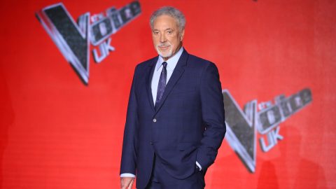Older man in a dark suit posing on a bright red backdrop with event graphics nearby, looking at the camera.
