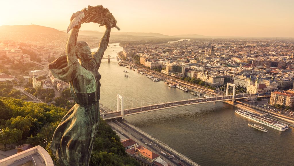 Bronze statue with arms raised overlooking Budapest’s Danube River at sunset, major bridges and city below.