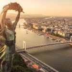 Bronze statue with arms raised overlooking Budapest’s Danube River at sunset, major bridges and city below.