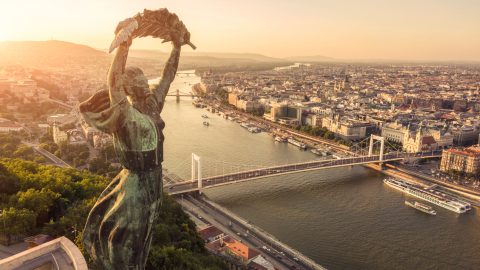 Bronze statue with arms raised overlooking Budapest’s Danube River at sunset, major bridges and city below.