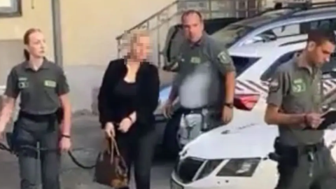 Woman in a black top being escorted by three uniformed officers beside a white police car in a parking area (face blurred).