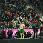 Hammer thrower in green shorts and white jersey inside a netted throwing cage, raising his right arm as photographers and a cheering crowd watch in the stands.