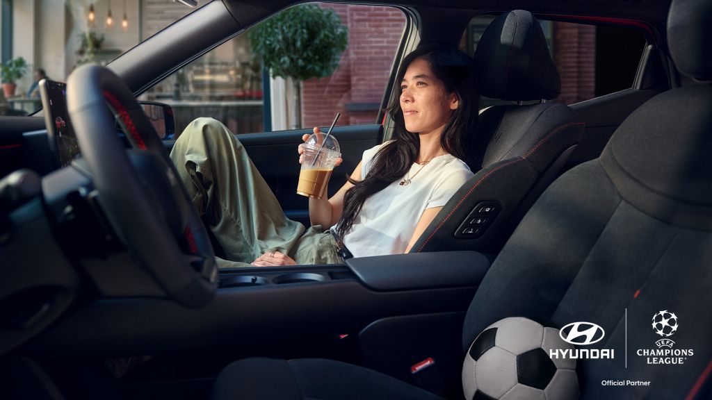 Woman relaxing in a car with an iced coffee, passenger soccer ball in view and logos for Hyundai and UEFA Champions League.