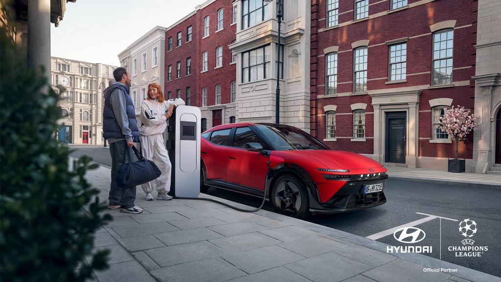 Red Hyundai electric car charging at a curbside station on a city street as two people chat nearby; logos visible in the corner.