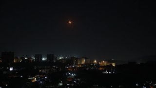Night city skyline with lit buildings; a small orange flare in the sky above.