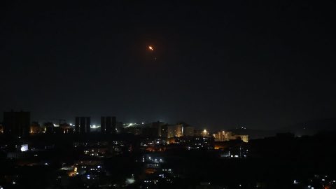 Night city skyline with lit buildings; a small orange flare in the sky above.
