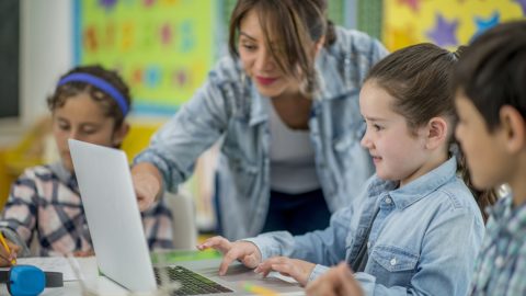 Teacher assists students using a laptop in a colorful classroom setting.