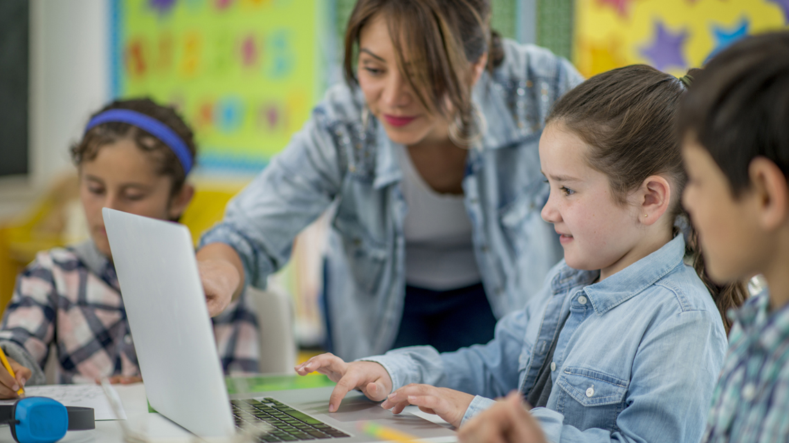 Teacher assists students using a laptop in a colorful classroom setting.