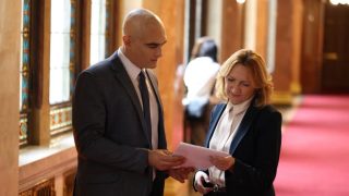 Two business professionals in a formal hallway review a document together, dressed in suits and white shirts.