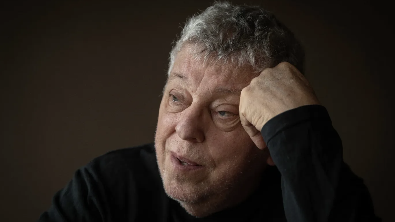 Older man with gray hair rests his head on his hand, looking contemplative in a dimly lit portrait.