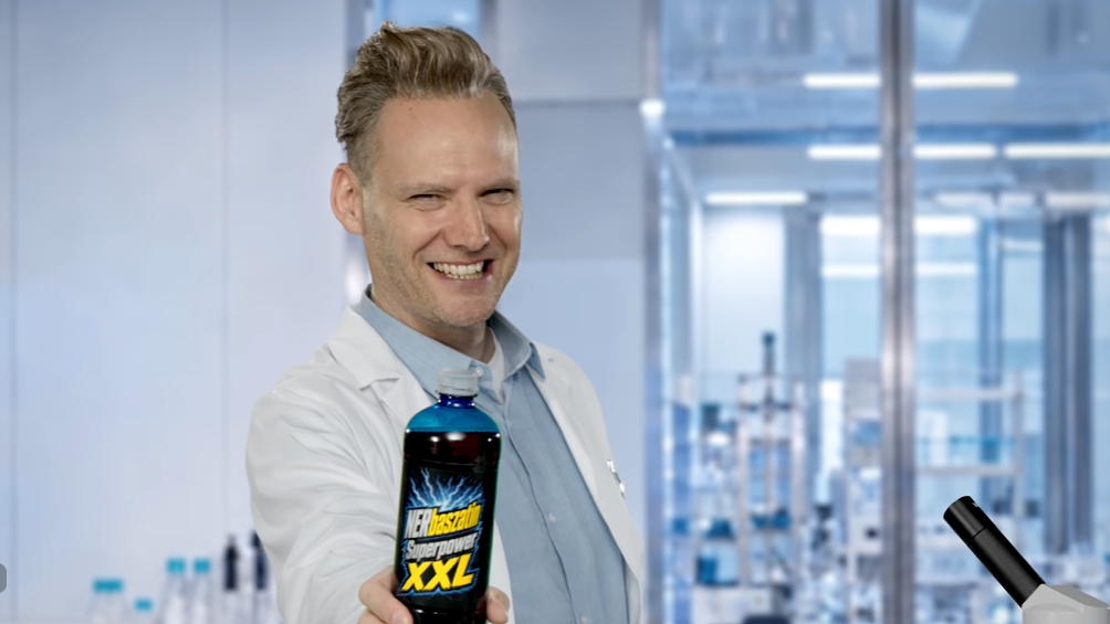 Smiling scientist in a lab coat extends a blue bottle labeled XXL toward the camera in a modern lab setting.