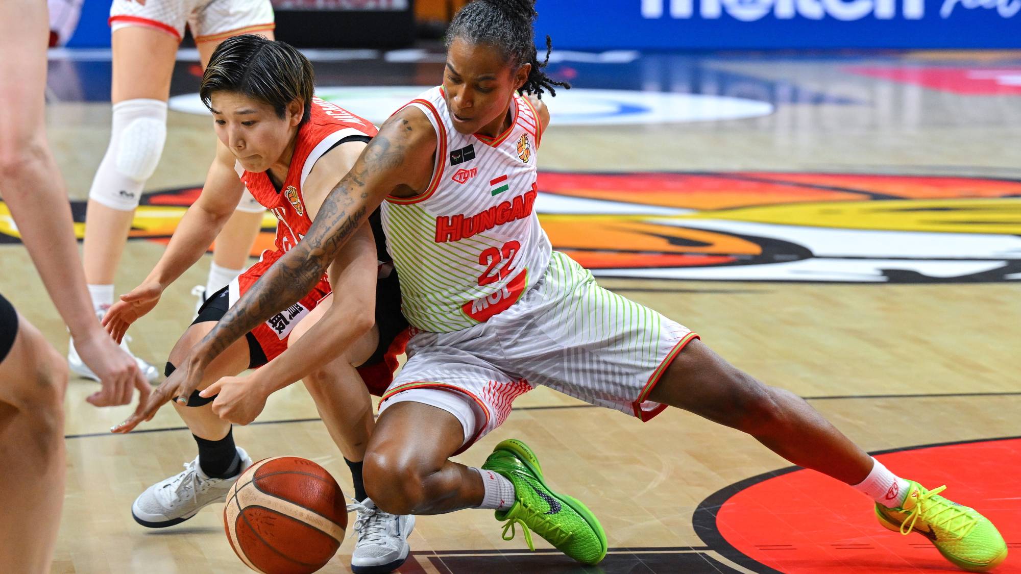 Two female basketball players reach for the ball during a fast-paced game on a wood court, with a Hungary jersey visible on one player.
