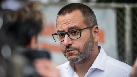 Close-up portrait of a man with short hair and glasses in a white shirt, looking off to the side outdoors.
